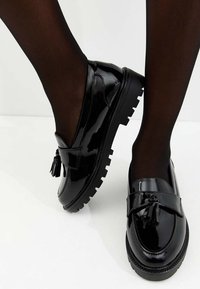 Mocassins en cuir verni noir avec une semelle épaisse, un détail à pompons et des accents texturés, présentés avec des collants noirs transparents.