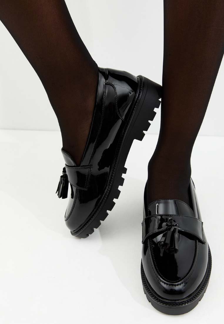 Mocassins en cuir verni noir avec une semelle épaisse, un détail à pompons et des accents texturés, présentés avec des collants noirs transparents.