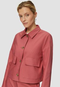 Roze jas gemaakt van lichtgewicht stof, met een kraag, vier grote voorzakken en ronde knopen. Gladde textuur met minimale naden.
