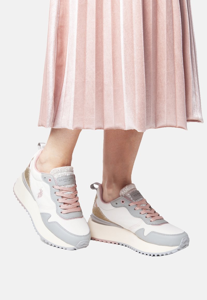 White and light pink sneakers with metallic accents, textured mesh, and a thick sole, paired with a pleated pink skirt.