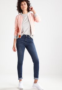 Giacca bomber rosa chiaro con chiusura a zip, t-shirt grigia, jeans in denim scuro e sneakers bianche, con i polsini dei jeans arrotolati.