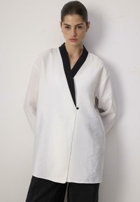 White button-up top with a black collar, featuring a textured fabric and a loose fit. Notable asymmetrical design and v-neckline.