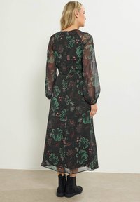 Bloemige midi-jurk met lange doorschijnende mouwen, een zwarte basis, groene en roze bloemenpatronen en een getailleerde taille. Zwarte enkelboots maken de look compleet.