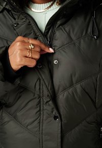 Donkergroene gewatteerde pufferjack met een ritssluiting aan de voorkant, drukknopen en zwarte manchetten. De hand is op de ritssluiting geplaatst.