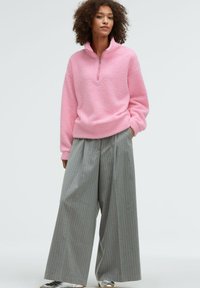 Hellrosa Fleece-Pullover mit Reißverschluss und texturierter Oberfläche, kombiniert mit grauen, gestreiften weiten Hosen. Silberschuhe runden das Outfit ab.