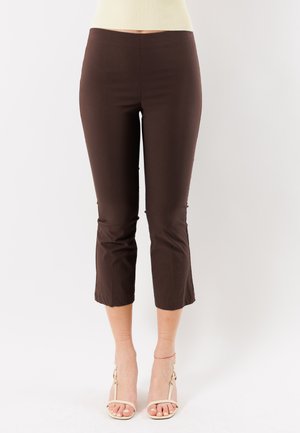 Donna con pantaloni aderenti marrone scuro accorciati e sandali crema con tacco, in piedi davanti a uno sfondo bianco uniforme.