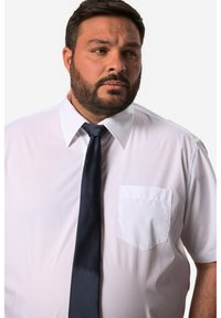 Camicia bianca con taschino sul petto, colletto classico e maniche corte, abbinata a una cravatta blu scuro. Tessuto dalla texture liscia.