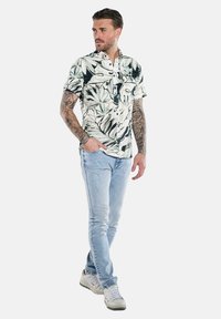 Kurzarmhemd mit einem tropischen Blattmuster in Grün und Schwarz, kombiniert mit hellblauen Jeans und weißen Turnschuhen.