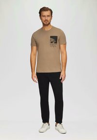 Beige Baumwoll-T-Shirt mit einem schwarzen grafischen Etikett, das ein Mondmotiv und Text zeigt, kombiniert mit schwarzen Hosen und weißen Sneakern.