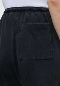 Μαύρο μαλακό υφασμάτινο παντελόνι με ελαστική μέση και τετράγωνη πίσω τσέπη, φορεμένο με λευκό πουκάμισο μαζεμένο μέσα.