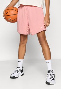 Ροζ σορτς μπάσκετ Nike με υφή διχτυωτού υφάσματος, σε συνδυασμό με μαύρα και λευκά παπούτσια. Μία μπάλα μπάσκετ κρατιέται στο αριστερό χέρι.