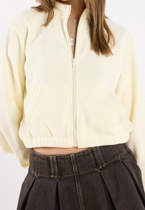 Vrouw draagt een korte crèmekleurige jas met ritssluiting aan de voorkant en zakken, gecombineerd met een hooggetailleerde donkerblauwe geplooide denimrok, waarbij gedeeltelijk de buik zichtbaar is.