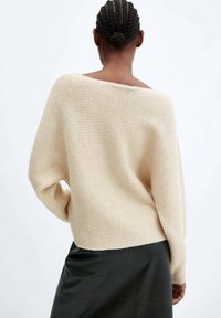 Pull en maille beige surdimensionné avec un large col et une texture côtelée. Porté avec une jupe en cuir noire, affichant un ajustement décontracté.