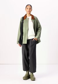 Jachetă verde cu guler maro, pulover alb, pantaloni evazați gri și cizme verzi. Modelul stă în picioare, ținând o geantă neagră.