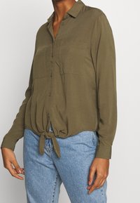 Oliivinvihreä, kevyt napillinen paita, jossa on etusolmuke ja kaksi rintataskua, pitkät hihat ja kaulus.