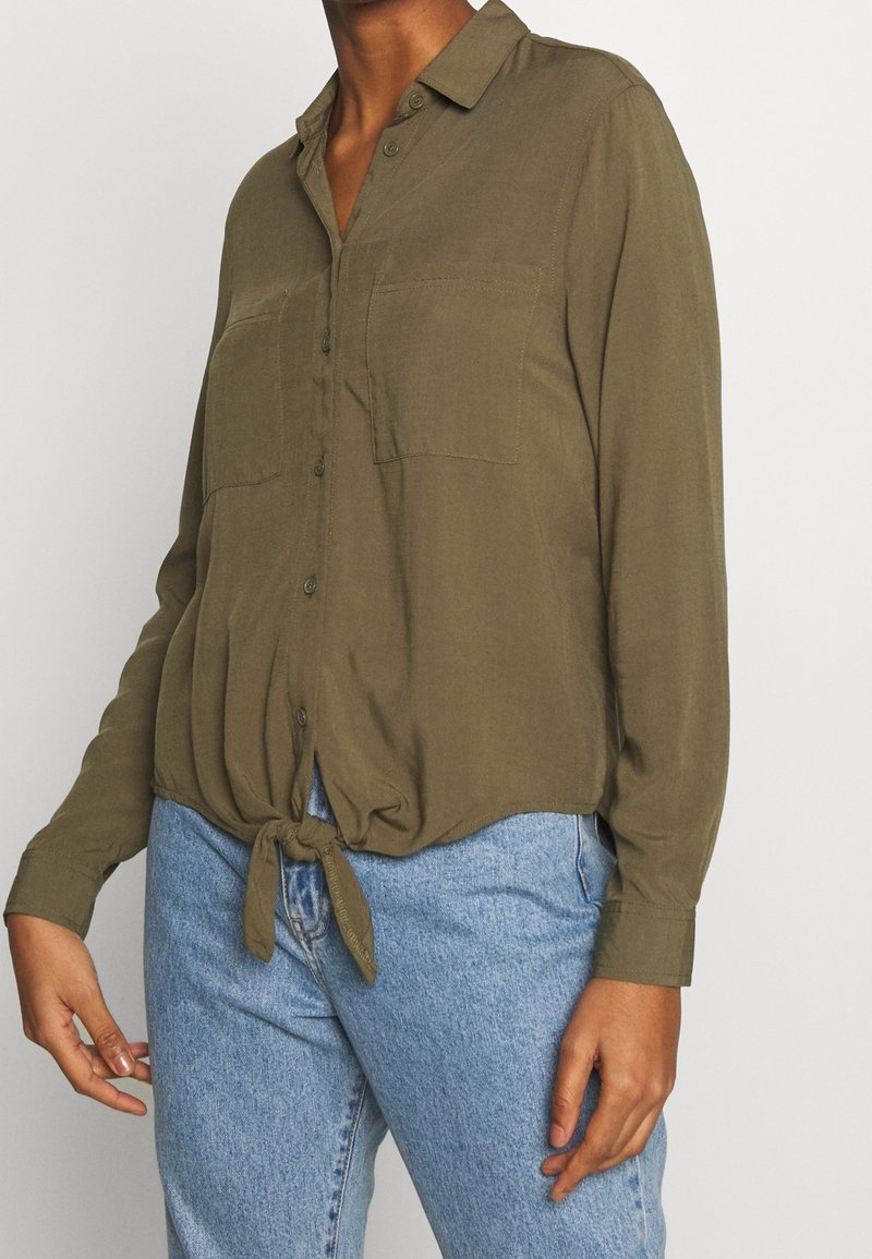 Oliivinvihreä, kevyt napillinen paita, jossa on etusolmuke ja kaksi rintataskua, pitkät hihat ja kaulus.