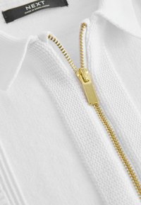 Maglione bianco a maglia con zip dorata, caratterizzato da un colletto a polo e un tessuto texture. L'etichetta mostra il nome del marchio "NEXT" e l'origine "Made in Bangladesh."