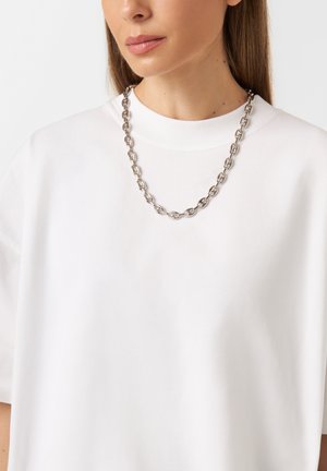 Collana in argento con maglie ovali, indossata sopra una maglietta bianca semplice con scollo rotondo, texture liscia e vestibilità rilassata.