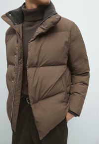 Braune Steppjacke mit hohem Kragen, ausgestattet mit einem grauen Fleece-Futter und einem Reißverschluss vorne. Sie hat gesteppten Paneele und Druckknöpfe.