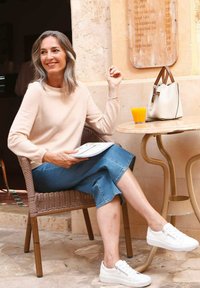 Lichtbeige trui, denim culottes, witte sneakers met ritsen. Geplaatst aan een tafel met een gestructureerd stenen oppervlak, een glas sinaasappelsap in de buurt.