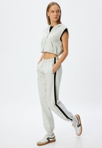 Grijze cropped hoodie met capuchon en rits; gecombineerd met grijze joggingsbroek met zwarte zijkanten. Model draagt witte sneakers.