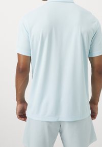 Jasnoniebieska koszulka polo z krótkimi rękawami, o miękkiej, gładkiej teksturze i standardowym kołnierzyku. Widok prezentuje tył koszulki.