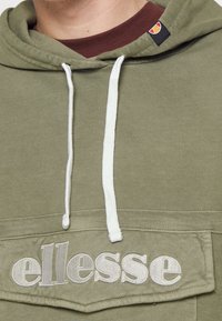Sudadera color verde oliva con cordones blancos, que presenta un logo bordado de "ellesse" en el bolsillo frontal y una capa interior en contraste de color burdeos.