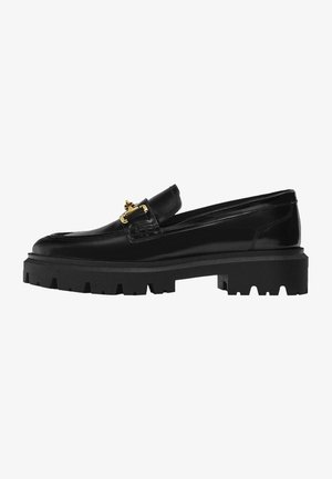 Mocassin en cuir noir avec semelle épaisse à crampons et détail boucle dorée sur le dessus.