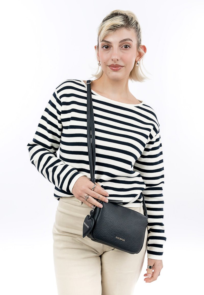 Junge Frau trägt schwarzen und weißen gestreiften Pullover und beige Hose, hält eine schwarze Leder-Umhängetasche mit herzförmigem Anhänger.