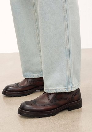 Zapatos brogue de cuero marrón oscuro con perforaciones decorativas, punta redondeada y suela de goma negra gruesa, combinados con denim azul claro.