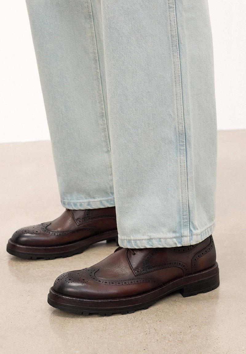 Scarpe brogue in pelle marrone scuro con perforazioni decorative, punta arrotondata e suola in gomma nera spessa, indossate con jeans di denim azzurro chiaro.