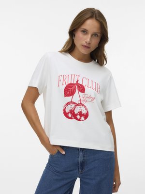 Νεαρή γυναίκα φοράει λευκό T-shirt με κόκκινες κερασιές και το κείμενο "FRUIT CLUB", συνδυασμένο με μπλε τζιν, στέκεται με ένα χέρι στην τσέπη.