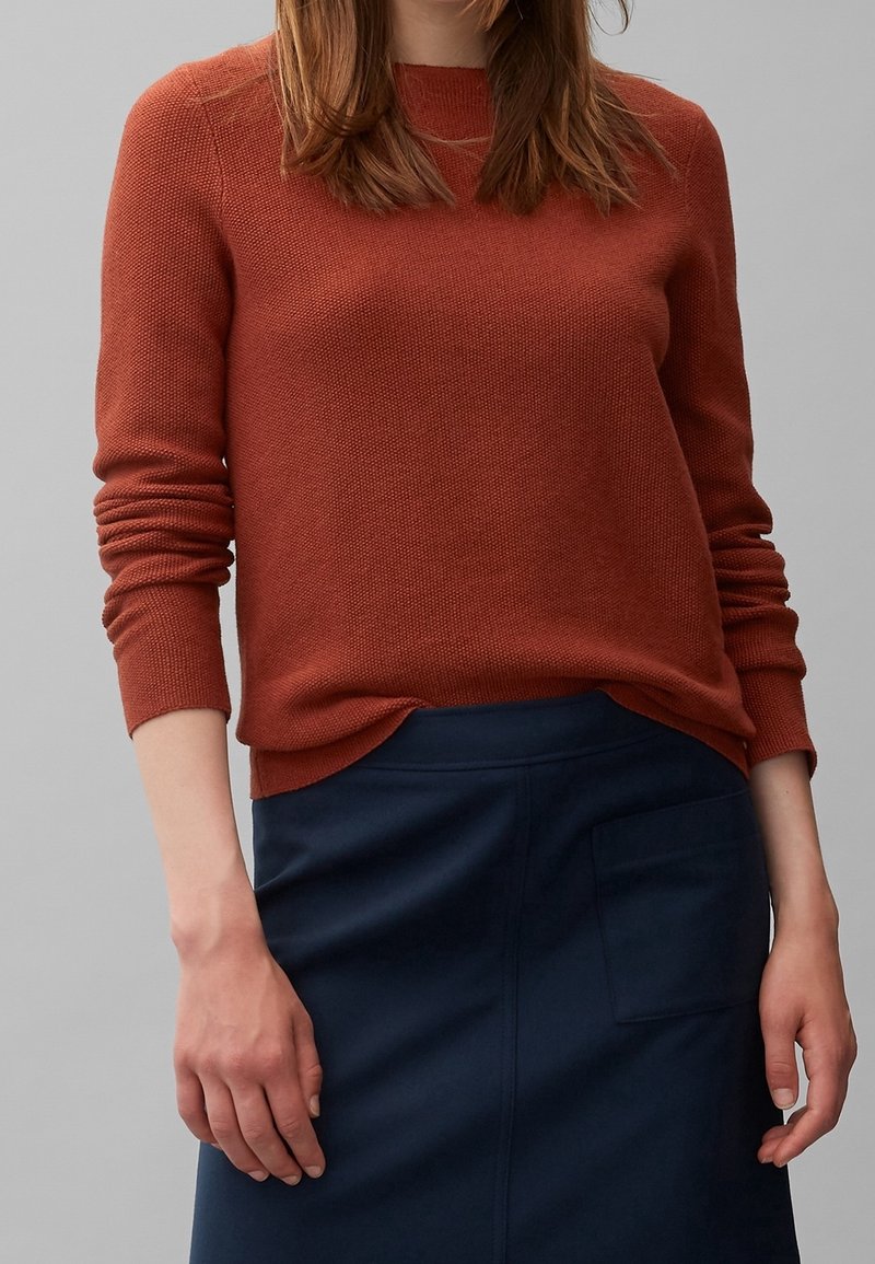 Strickpullover in Rostfarbe mit langen Ärmeln, Rundhalsausschnitt und geripptem Saum, kombiniert mit einem dunkelblauen Rock mit Seitentasche.
