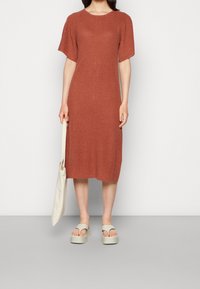 Ein rostfarbendes knielanges Kleid mit kurzen Ärmeln, gerippter Textur, zentralseitiger Nahtdetail, getragen mit hellen Sandalen und einer beigen Tragetasche.