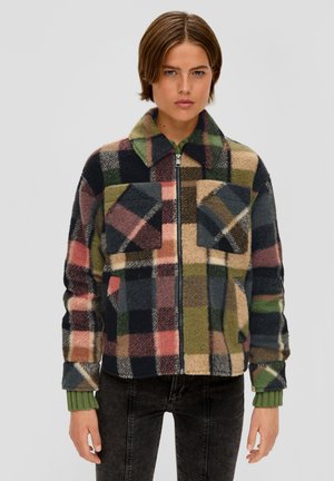 Jongere die een multikleurige geruite zip-up jas draagt met twee borstzakken, groene gebreide manchetten en zwarte jeans tegen een effen achtergrond.