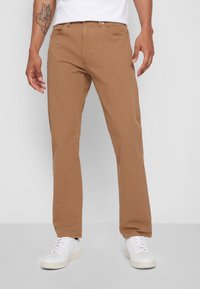 Braune Baumwoll-Hose mit geradem Bein und glatter Textur, zwei Vordertaschen und einem Knopfverschluss, kombiniert mit weißen Turnschuhen.