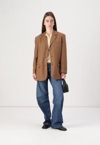 Brun overdreven blazer med spiss lapel, beige bluse, vide jeans og svart håndveske. Glatt stoff, avslappet passform, minimalistisk design.