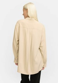 Beige Langarmhemd mit entspanntem Schnitt, weichem Stoff, Knopfmanschetten und Rückenfalte. Das Design verfügt über einen abgerundeten Saum.