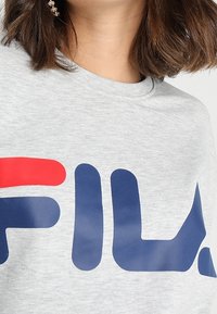 Gray sweatshirt featuring a large "FILA" logo in red and navy blue. Soft fabric, crew neckline, and relaxed fit with short sleeves.