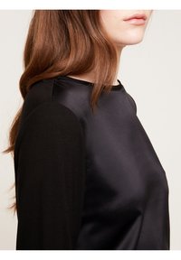 Svart topp med en slät satinframsida och texturerade ärmar. Designen har en rund halsringning och mjuka draperingar längs sidorna.