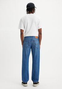 T-shirt bianco a maniche corte, jeans in denim blu a gamba larga con una toppa in pelle marrone e cappellino nero. Tasche posteriori visibili con dettagli di cucitura.