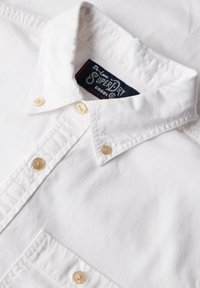 Witte overhemden met een licht gestructureerde stof, puntige kraag en vier beige knopen. Bevat een blauw label met de tekst "Superdry."