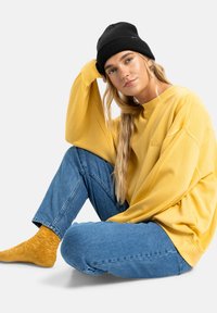 Geel oversized sweatshirt, blauwe denim jeans en een zwarte gebreide beanie. Model zit met lang haar en toont een zachte, textuur sok.