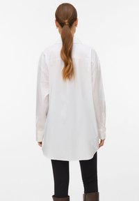 Hvit langermet skjorte med knappelukking foran, buet kant og avslappet passform. Materialet virker glatt; har knappemansjetter og krage.