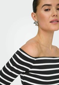 Sølvblomsterformede øreringe har lagdelte petaldesigns. Modellen bærer en sort-hvid stribet off-shoulder top med en ribbet tekstur.