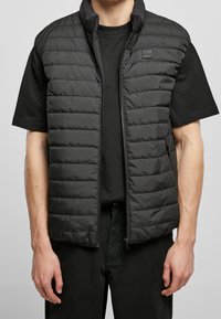 Černá polstrovaná vesta s vysokým límcem a zapínáním na zip, se čtvercovou logem nášivkou. Nosená přes černé tričko s krátkým rukávem.