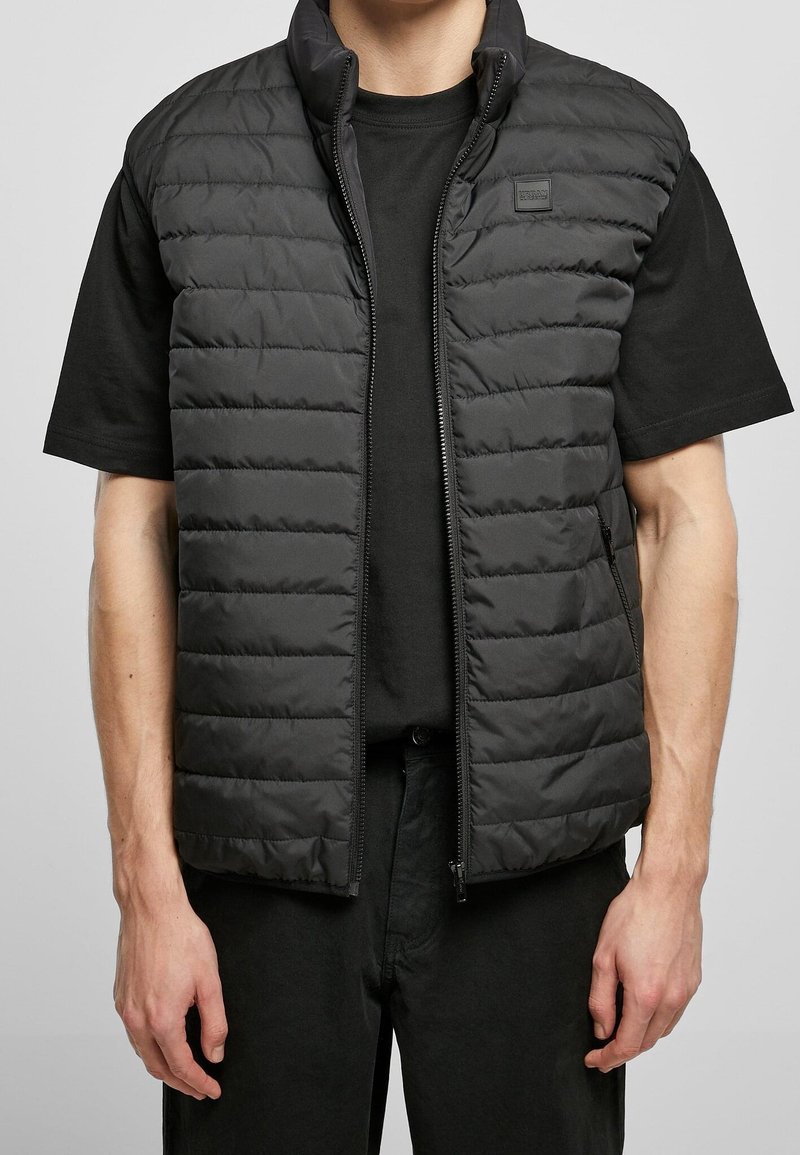 Černá polstrovaná vesta s vysokým límcem a zapínáním na zip, se čtvercovou logem nášivkou. Nosená přes černé tričko s krátkým rukávem.