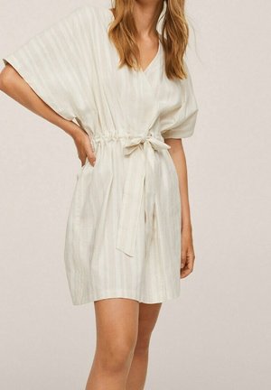 Vrouw draagt een korte, roomkleurige wikkeljurk met brede korte mouwen en een gestrikte tailleband, staand tegen een neutrale achtergrond.