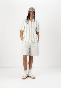 Lichtgroen shirt met korte mouwen en een patroon, gecombineerd met lichtbeige shorts. Het model draagt een grijze pet en witte sneakers.