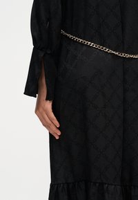 Robe noire avec motif texturé et ceinture chaîne. Comprend des manches longues avec détail fendu et ourlet superposé à volants en bas.